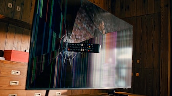 Primer plano de un televisor con la pantalla rota y líneas de colores, con un control remoto negro incrustado. La pantalla muestra un partido de fútbol con un jugador y público de fondo