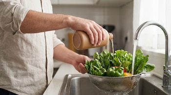 Primer plano de una persona lavando vegetales en un colador de metal bajo el grifo de la cocina, vertiendo líquido de una botella marrón sobre ellos.