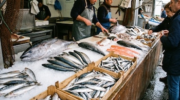 Puesto de mercado con pescado fresco sobre hielo: atún, caballa, sardina y lomos de salmón. Tres hombres con delantal y dos clientes observan el producto.