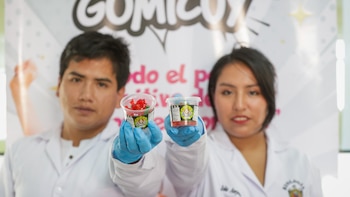 Dos jóvenes en batas de laboratorio y guantes azules sostienen envases transparentes con gomitas rojas y naranjas llamadas GOMICUY frente a un logo