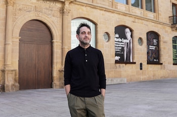 Hombre con polo oscuro y pantalones verdes posa ante una fachada de piedra con un arco de madera, pilares y dos carteles de exposición visibles