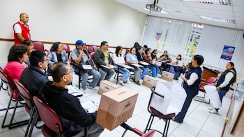 Un grupo de personas sentadas en semicírculo escucha a dos instructoras junto a cajas de cartón marcadas con la sigla ONPE
