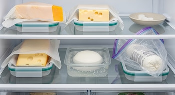 Interior de una heladera con quesos variados en recipientes y papel, una bola de mozzarella en agua y una pequeña fuente con cubos blancos.