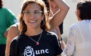 Primer plano de una mujer sonriente con gafas, aros, collar y camiseta negra con el logo y las letras 'unc'. Personas borrosas en el fondo