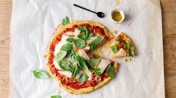Vista aérea de una pizza con una porción separada, cubierta con salsa de tomate, mozzarella, albahaca fresca, cebolla roja y parmesano, sobre papel de horno blanco.