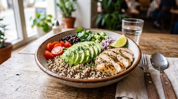Un plato hondo sobre una mesa contiene quinoa, láminas de palta, pollo asado, tomates cortados, frijoles negros y cubiertos al lado.