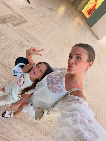 Cami Homs y una niña posan para una selfie en un suelo de baldosas claras. Cami viste de blanco; la niña, con camiseta y shorts, saca la lengua y hace la señal de la paz