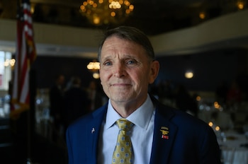 Retrato de Ed Gallrein, un hombre de mediana edad con traje azul, camisa celeste y corbata dorada, sonriendo ligeramente, con la bandera de EE.UU. al fondo