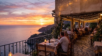 Mesas dispuestas en la terraza de un restaurante con personas cenando bajo luces decorativas frente al mar durante el atardecer.