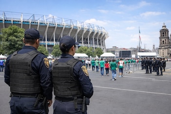 Dos agentes de policía, vistos de espalda con chalecos antibalas, supervisan una gran multitud caminando frente a un estadio y una catedral bajo un cielo azul.