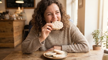 Mujer con cabello rizado y suéter beige comiendo una galleta de arroz. Hay migas en su cara y un plato con más galletas sobre una mesa de madera.