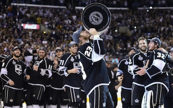 Un jugador de hockey sobre hielo levanta y besa un trofeo metálico rodeado por sus compañeros de equipo vestidos con uniforme negro