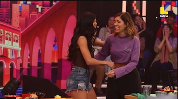 Korina Rivadeneira y Paloma Fuiza se abrazan y ríen en un set de televisión, frente a una audiencia borrosa y un fondo animado de ciudad roja