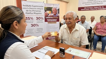 Una mujer entrega la Tarjeta Dorada a un hombre mayor sonriente. En el escritorio hay documentos. Banners promocionales del programa de Guerrero en el fondo.