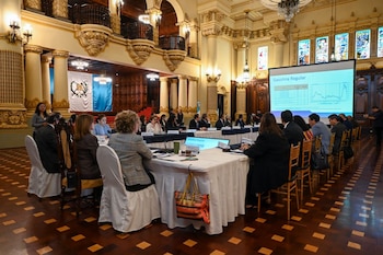 Salón ornamentado con personas sentadas alrededor de una mesa larga; pantalla de proyección muestra gráficos de gasolina regular; banderas de Guatemala en la pared