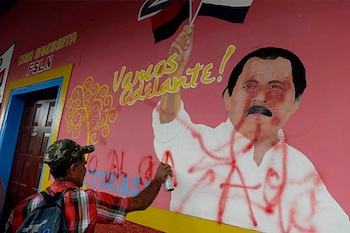Un hombre de espaldas, con gorra camuflada y camisa a cuadros, rocía pintura roja sobre un mural rosado del rostro de Daniel Ortega