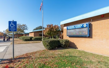 Vista exterior de la Escuela Primaria Isador Cohen con letrero digital de conferencias, mástil con bandera, cartel de ruta accesible y paredes de ladrillo