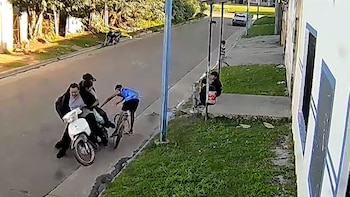 Dos hombres en motocicleta interceptan a un joven en bicicleta en una calle. Al fondo, se ven árboles, edificios y personas sentadas en una parada de autobús