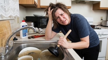 Mujer con expresión de frustración, pelo revuelto, sosteniendo un desatascador en un fregadero de cocina lleno de agua sucia y platos apilados.