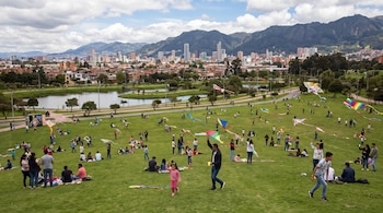 Un gran parque verde con muchas personas volando cometas de colores; al fondo se ven edificios de una ciudad y montañas bajo un cielo nublado.