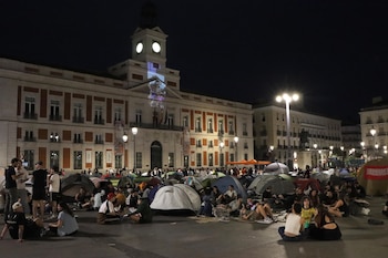 Grupo de personas sentadas y acostadas entre tiendas de campaña en una plaza iluminada de noche frente a un edificio con reloj