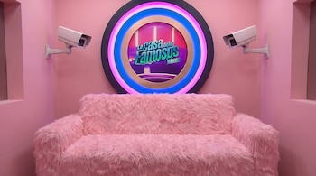 Un cuarto rosa con un sillón de peluche rosa, dos cámaras de seguridad blancas y un logo circular luminoso de La Casa de los Famosos México.