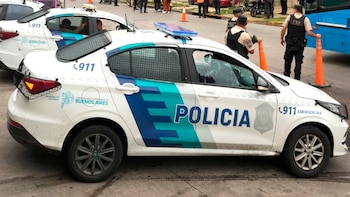 Patrullero blanco de la Policía de Buenos Aires con franjas azules y logo. Ventanas traseras con rejas. Policías y conos de seguridad en el fondo