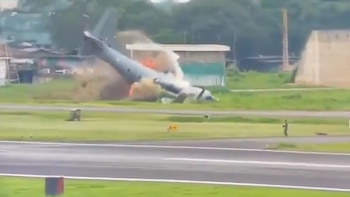 Un avión militar gris destrozado y en llamas, con denso humo elevándose, yace en un campo verde junto a una pista de aterrizaje. Hay un edificio al fondo