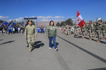 Keiko Fujimori y un militar caminan en una carretera frente a una banda militar, soldados con bandera de Perú y escaleras de avión bajo un cielo azul