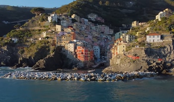 Vista aérea de un pueblo costero con casas de colores brillantes en la ladera de una montaña rocosa, el mar azul en primer plano y un rompeolas de rocas