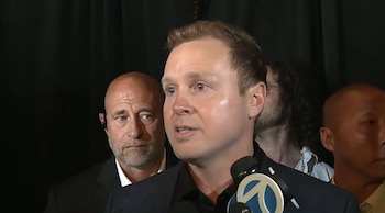 Spencer Pratt, un hombre caucásico de cabello claro, habla ante un micrófono con el logo de un canal de noticias, rodeado por otras personas