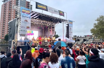 Un escenario exterior con una banda de músicos tocando instrumentos frente a una multitud. Hay banners publicitarios, edificios y árboles al fondo