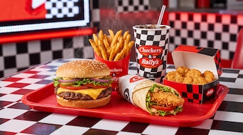 Una hamburguesa con queso, un enrollado de pollo, papas fritas, pollo frito y una bebida sobre una bandeja roja en una mesa de azulejos a cuadros.