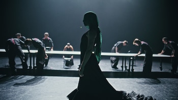 Una mujer con un velo verde y vestido oscuro se para en un escenario oscuro. Otros artistas en ropa oscura se inclinan sobre mesas largas en el fondo