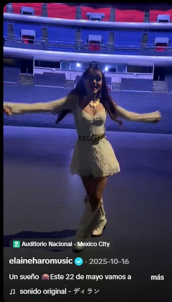 Mujer joven de cabello oscuro con vestido claro, cinturón, collar y botas vaqueras, brazos extendidos, en un escenario con asientos de estadio al fondo