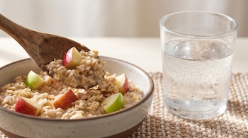 Una cuchara de madera saca avena con trozos de manzana de un bol. Un vaso de agua con gotas de condensación está a un lado.