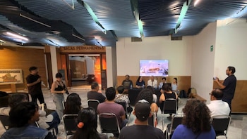 Salón con techo acanalado, luminarias y público sentado. Un panel de cinco personas frente a una pantalla. Un hombre de pie con micrófono y otro con móvil