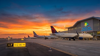 Pista del Aeropuerto Internacional de Punta Cana al atardecer. Aviones comerciales en fila y uno aterrizando, con terminal moderna y su logo bajo un cielo naranja y azul.