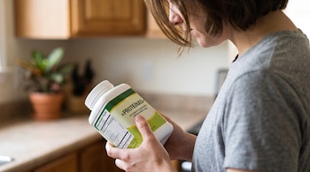 Una persona sostiene un envase blanco con etiqueta nutricional de suplemento de proteínas en una cocina con muebles de madera.