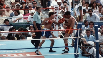 Dos boxeadores con guantes rojos pelean en un cuadrilátero rodeado por un árbitro, fotógrafos y público en las graderías