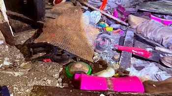 Varios trabajadores con cascos amarillos y chaquetas naranjas empujan una pared de ladrillo entre escombros y vegetación