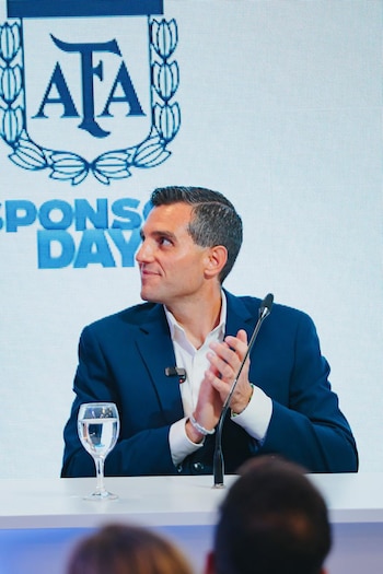Hombre de traje azul y camisa blanca sentado en una mesa, aplaudiendo. Detrás, una pantalla con el logo de AFA y texto "SPONSOR DAY". Hay un vaso de agua y un micrófono en la mesa