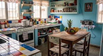 Cocina con paredes azules, azulejos de colores. Estufa, fregadero, vajilla en estantes, mesa de madera y sillas. Frutero con piña, plátanos y frutas. Radio.