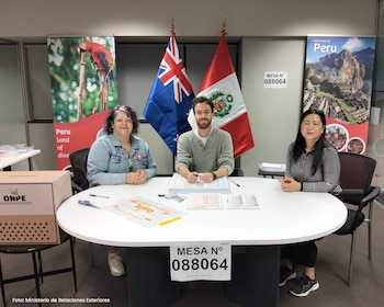 Tres personas sentadas en una mesa frente a banderas de Nueva Zelanda y Perú. Hay una urna electoral con el logo ONPE y pancartas turísticas