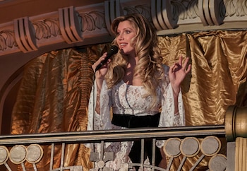 Mujer rubia con micrófono, vestido de encaje blanco y cinturón negro, sobre un balcón con barandilla metálica frente a cortinas doradas y detalles arquitectónicos