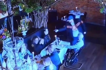 Toma de cámara de seguridad en un restaurante donde un hombre con gorra azul apunta con un arma corta hacia personas sentadas a una mesa