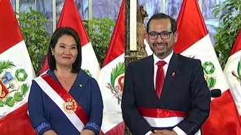 Dos personas de pie, sonrientes, con bandas presidenciales rojiblancas sobre trajes oscuros, flanqueadas por banderas de Perú y plantas verdes