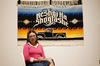 Una mujer joven con gafas y blusa rosa está sentada frente a un tapiz multicolor con el dibujo de una camioneta negra, texto blanco y patrones