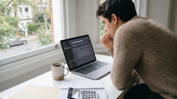Un hombre en suéter de lana mira atento una laptop en un escritorio blanco, junto a una taza de café, cuaderno y calculadora, con una ventana de fondo.