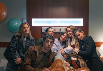 Siete personas, incluyendo un hombre y seis mujeres, sonríen juntas en una habitación de hospital con globos. Una mujer en pijama sostiene un gorro de bebé